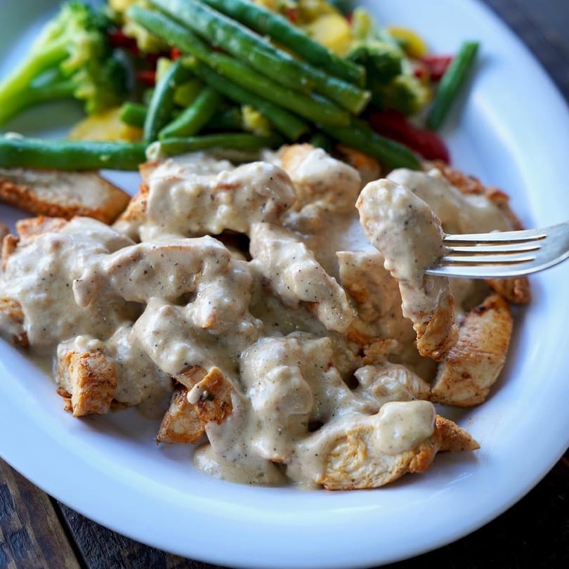 Creamy chicken pieces on a white plate with green beans and mixed vegetables.