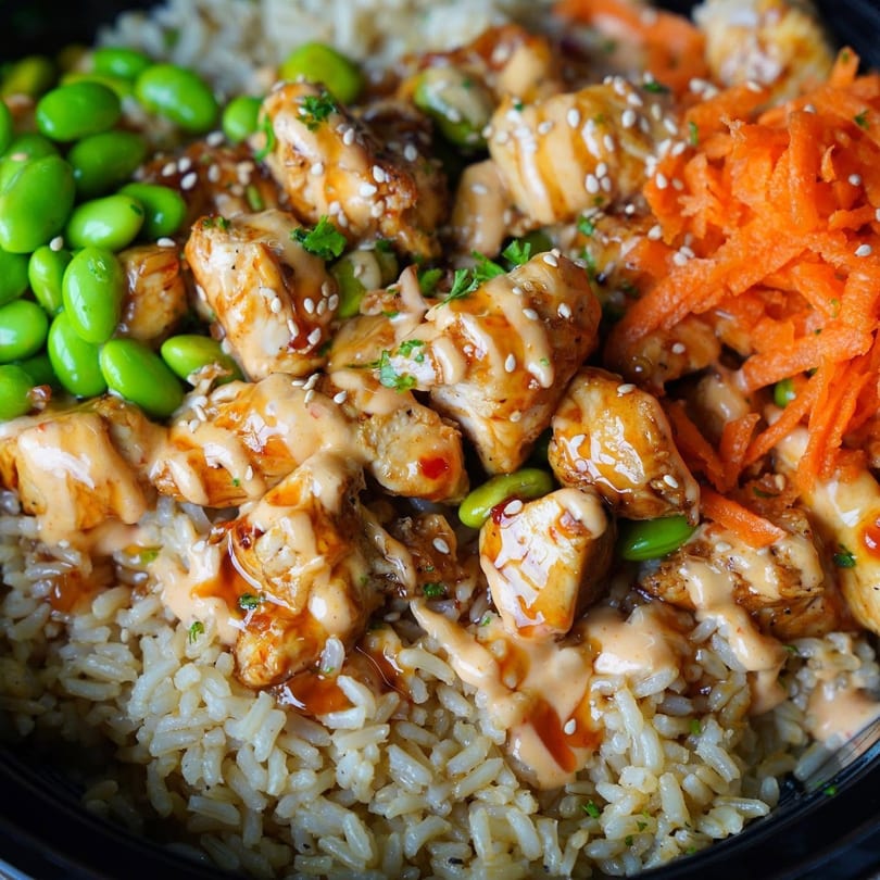 Close-up of a chicken and rice bowl with edamame, shredded carrots, and creamy sauce.